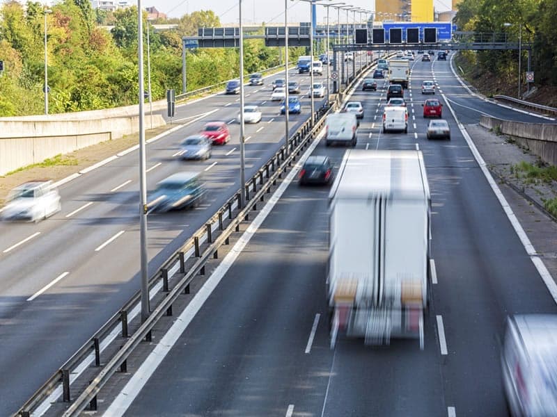 Drukke snelweg met meerdere rijstroken in beide richtingen, gefotografeerd vanuit een verhoogd standpunt. Personenwagens en een witte vrachtwagen rijden met hoge snelheid, zichtbaar als bewegingsonscherpte. Langs de weg staan verlichtingspalen en een matrixbord. Op de achtergrond zijn een viaduct en groen zichtbaar.