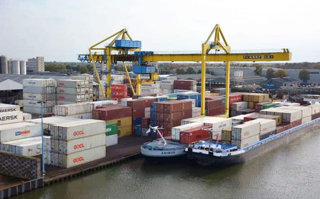 Luchtfoto van een binnenvaart containerterminal met het afgemeerde binnenvaartschip "Animar". Op de kade staan grote gele portaalkranen die containers overslaan tussen het schip en de opslagplaats. De kade ligt vol gestapelde zeecontainers van onder andere Maersk en OOCL. Op de achtergrond zijn bedrijfsgebouwen en bomen zichtbaar.