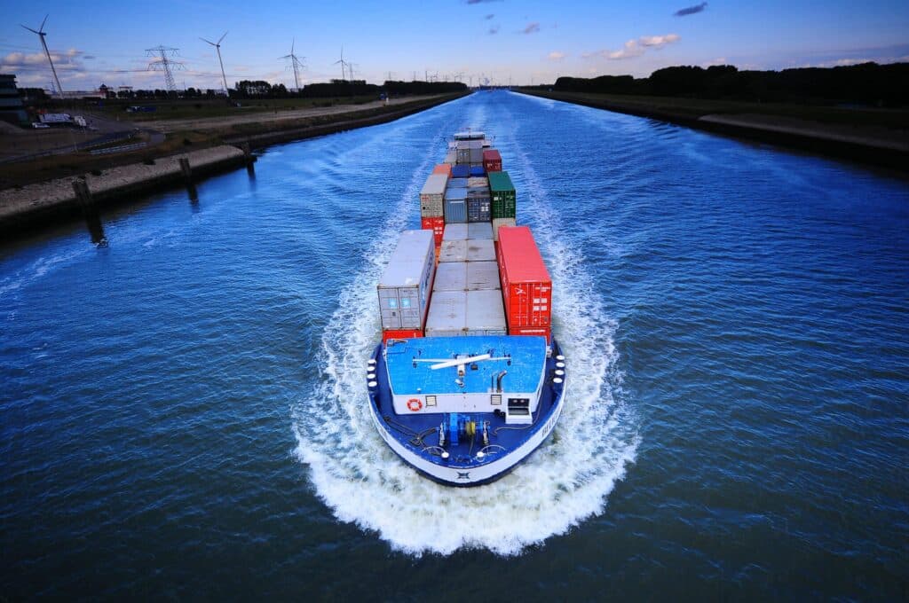 Een blauw binnenvaartschip beladen met gekleurde zeecontainers vaart over een rechte Nederlandse kanaal, gefotografeerd vanuit de lucht. Het schip trekt een brede kielzog aan beide zijden. Op de achtergrond zijn vlakke polders zichtbaar met windturbines en hoogspanningsmasten langs de oever.